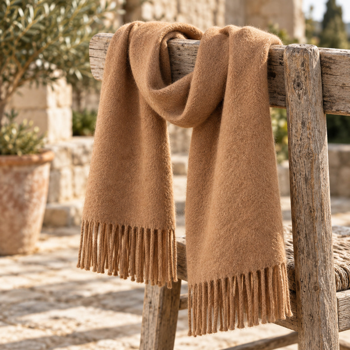 A soft, light brown scarf with fringed ends is draped over the backrest of a rustic wooden chair outdoors, with a stone patio and potted plant in the background.