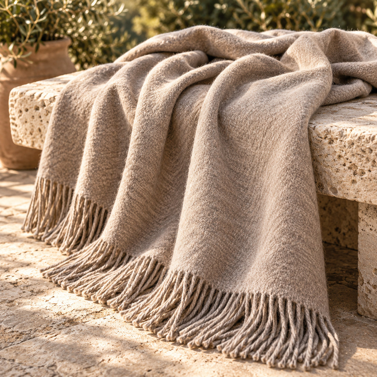 A beige, fringed wool throw blanket draped over a stone bench outdoors, with sunlight highlighting its texture and potted plants in the background.